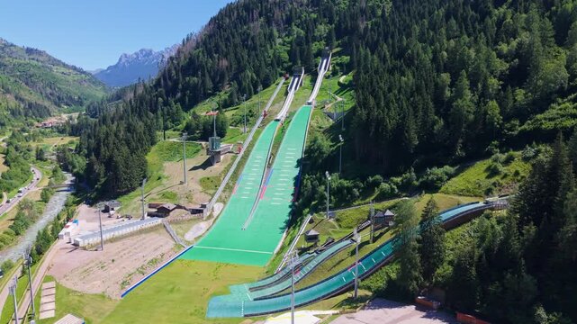 Aerial drone view of the predazzo ski jumping stadium in val di fiemme, surrounded by the italian dolomites, showing the summer ski jumping ramps and alpine landscape.