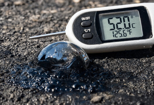 Digital thermometer measuring extreme heat on melting asphalt road surface.