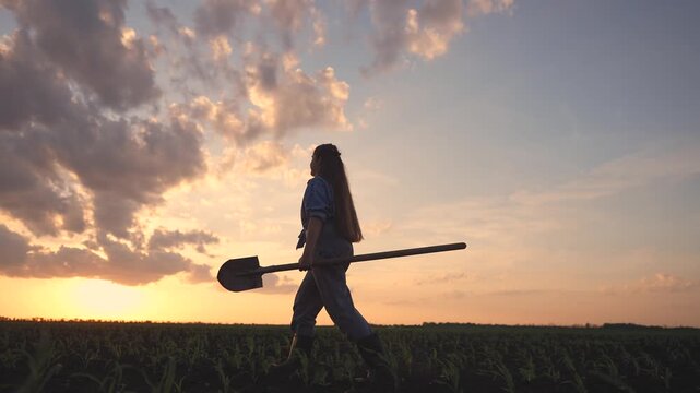 A teenage girl walks through a field with a shovel in hand, embodying the role of a teenage farmer during sunset. The teenage farmer takes in the beauty of the expansive farmland.