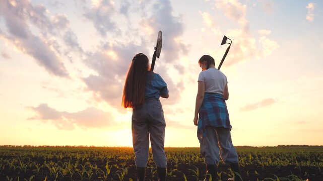 A girl and a woman stand together in a field, looking at the sunset. The girl holds a shovel while the woman carries a hoe, both wearing casual attire.