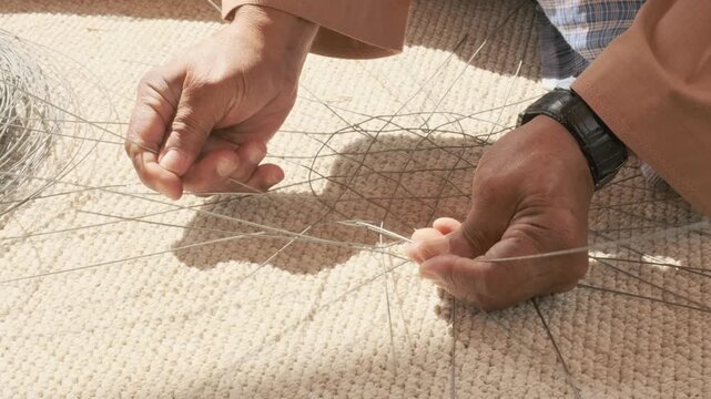 elderly man hands carefully twist and shape metal wire creating fish trap structure. ghazl al gargour shows authentic arabic craft. ghazl al gargour passes mastery down through generations.