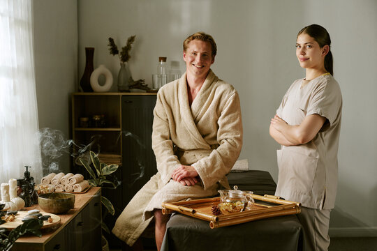 Portrait of smiling Caucasian young adult male client in bathrobe and female masseuse looking at camera in wellness spa. Image ideal for luxury resort marketing and beauty salon promotion