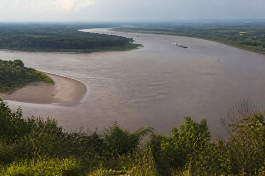 View from high bluff of wide brown river bending through low green forest, sandbar, tugboat and barge downstream, Mississippi River landscape