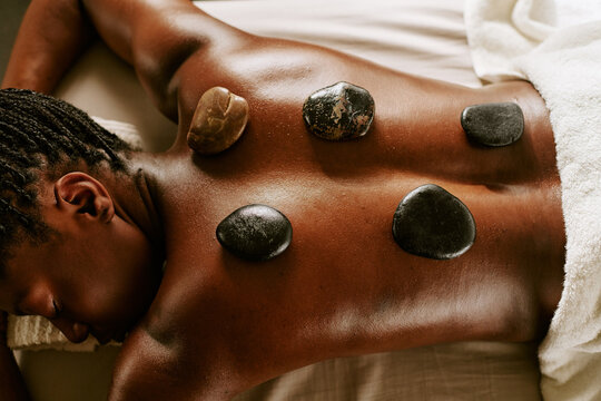 Young adult Black woman receiving hot stone massage therapy on back at wellness spa. High angle view of relaxing body treatment ideal for beauty salon advertising