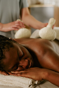 Vertical image of young adult African American woman receiving herbal compress massage on back at wellness spa, promoting holistic therapy and self care services
