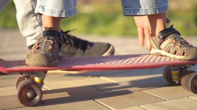 A boy practices balancing on a skateboard while standing on the skateboard. A man assists the boy by holding the skateboard for support.