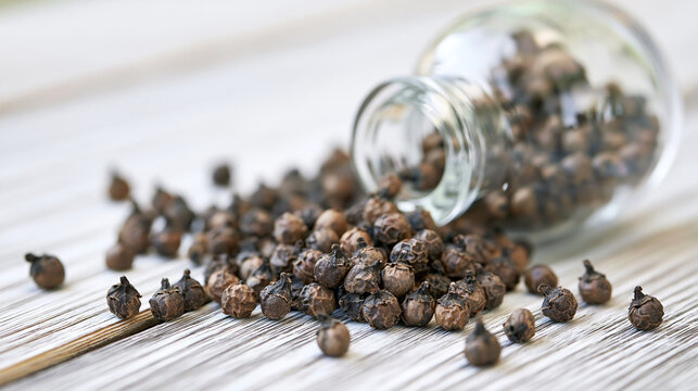 Black peppercorns spilling from a glass jar onto a light wooden table, representing seasoning, cooking, and culinary ingredients for food preparation