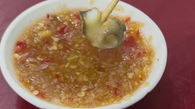 Close up of skewered spotted babylon snail dipping into and lifting from spicy chili ginger fish sauce with dripping sauce