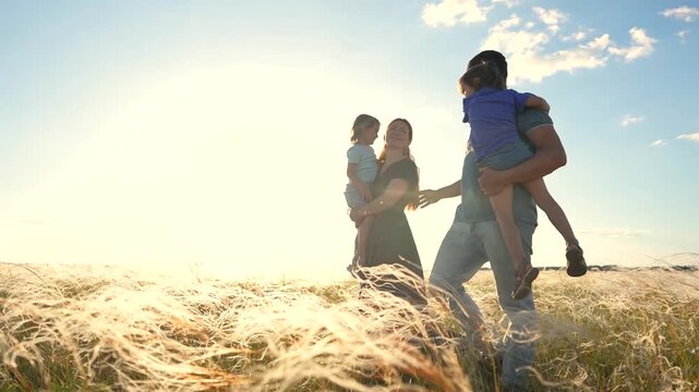 The woman and man enjoy time with their children in a field. In the field, the woman carries the girl, while the man holds the boy in the evening light.