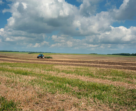 A blue wheeled tractor performing field work cultivation or plowing of the soil in a large open space under a cloudy sky