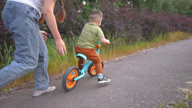 A woman helps a boy ride a blue bike as they move along a path. The boy, focused on biking, is assisted by the woman in a natural setting.
