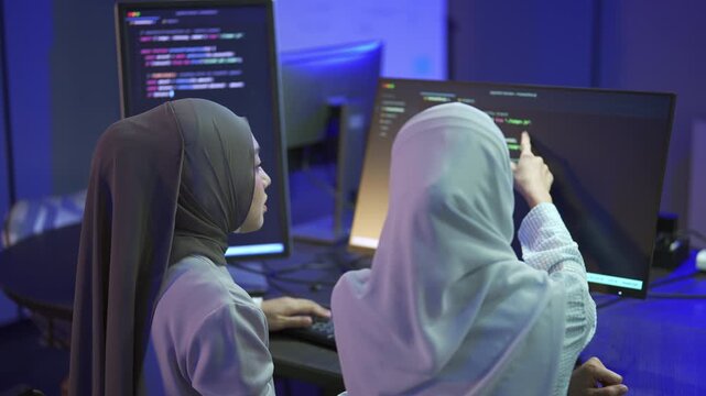 Rear view of Two Muslim women programmers in hijab collaborating on software code in a modern tech office.