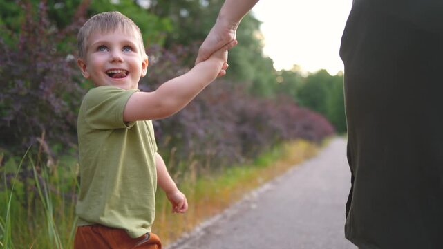 A woman holds a boy's hand as they walk along a path. The boy looks back, enjoying the nature around them and the woman s companionship.