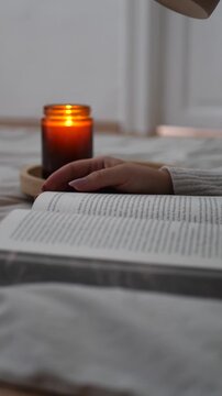 Hand resting on open book in bed with candle light and coffee in blurred background, low angle shot