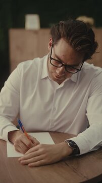 Vertical portrait of thoughtful businessman in glasses sitting at desk, writing on piece of paper, pausing to think, continuing with smile of satisfaction on face. Shooting in slow motion.