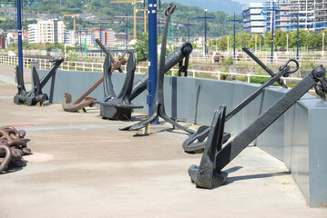 Fototapeta premium Row of historic ship anchors along a quay wall with tower cranes behind