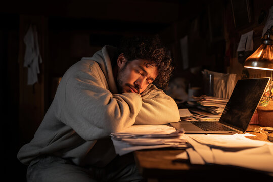 Man sitting alone near scattered papers, laptop, eyes closed, deep in exhaustion, indoor, dim light