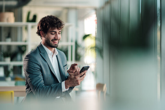 Young businessman using smartphone in modern office, smiling while checking messages or browsing internet on mobile