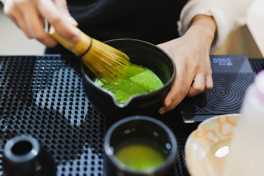 Barista's hand whisking green matcha powder using a traditional bamboo whisk.