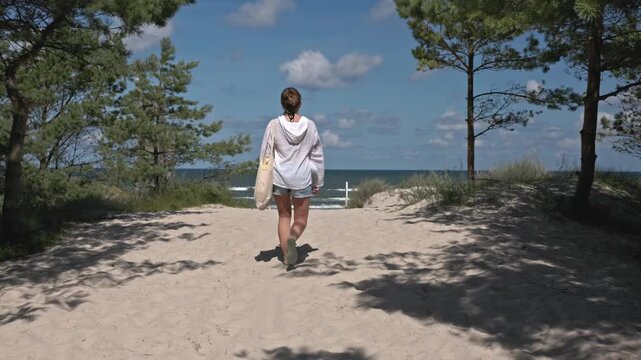 Woman in white hoodie walking along a sandy path through pine trees towards the Baltic Sea beach.