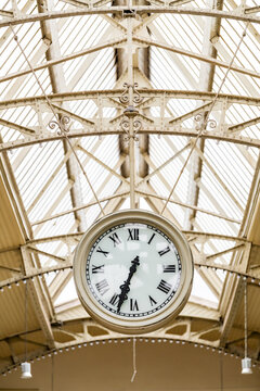 Large vintage clock hanging beneath an ornate glass and iron station roof. Historic architecture symbolizing travel, punctuality