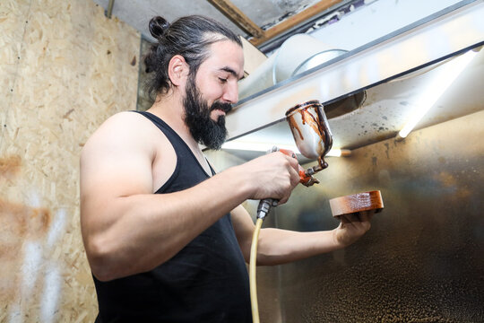 BEARDED MAN SPRAY COATING SMALL CYLINDRICAL PART IN WORKSHOP USING AIRBRUSH TOOL, WORKING AT INDUSTRIAL SPRAY STATION WITH PROTECTIVE VENTILATION, METAL SURFACES, WOOD PANEL BACKGROUND, SKILLED