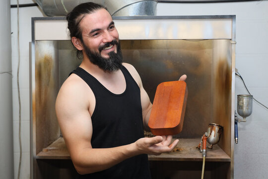 SMILING CRAFTSMAN HOLDING WOODEN BLOCK INSIDE INDUSTRIAL SPRAY BOOTH WITH METAL EQUIPMENT, SHOWING MANUAL WORK PROCESS, FINISHING STAGE, AND SMALL WORKSHOP ENVIRONMENT