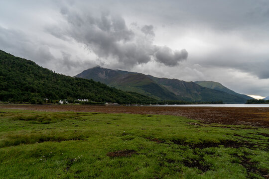 Scenic view of the dramatic coastal shore at Loch Leven with lush green and orange fields, tidal mudflats, and forested mountains rising under a stormy overcast sky in the Scottish Highlands