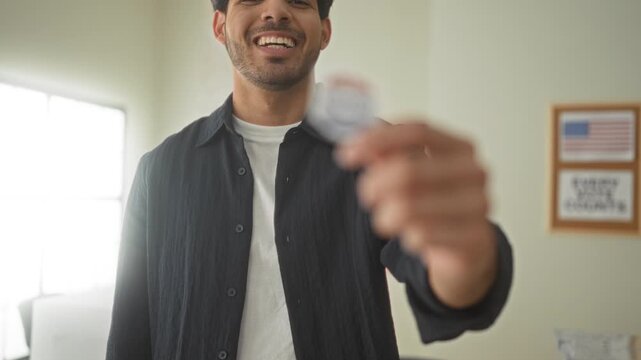 Young hispanic man holding an i voted sticker between thumb and forefinger, smiling and reaching forward with his hand in a polling building with flag poster visible; civic pride.