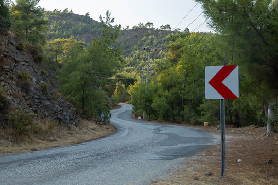 Winding asphalt road with a sharp curve warning sign passing through a lush green forest and rocky hills. Turkey, Mediterranean.