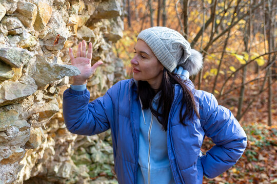 Adult woman pauses beside a weathered stone wall and studies its masonry during an autumn walk through the forest. Warm foliage and natural light create a travel image about historical discovery, aban