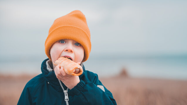 Child in Orange Beanie Eating Sausage Roll, Teal Jacket Outdoors