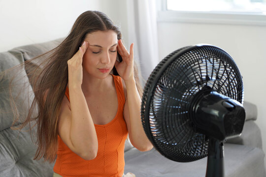 Young woman with a headache holds her temples in front of a running fan, suffering from the summer heat at home
