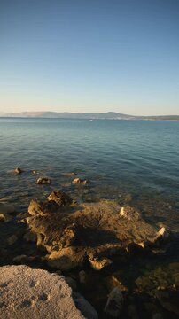 High-angle view of a rocky shoreline along clear Adriatic coastal waters illuminated by warm golden sunlight, with distant hills across the calm sea, 4K vertical video