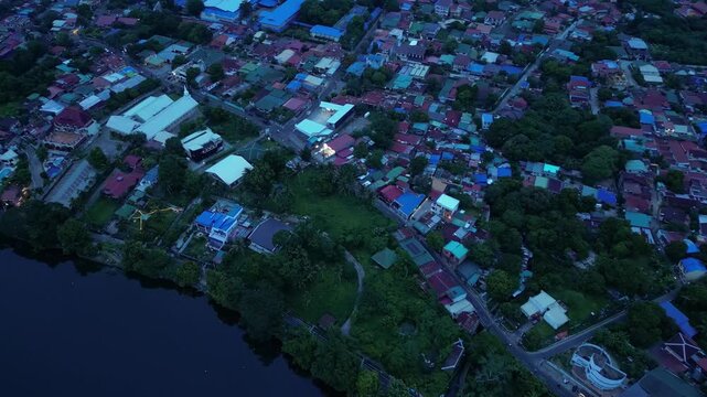 Slow cinematic drone footage flying above Sampaloc Lake, San Pablo City, Laguna, Philippines featuring calm freshwater, lakeside neighborhoods, tree-lined roads, and soft blue hour skies.