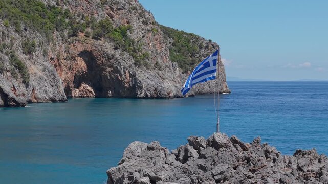 Drone orbit around a waving Greek flag, gradually revealing the crystal-clear waters, golden shoreline, and spectacular coastal landscape of Limnionas Beach on Evia Island, Greece.