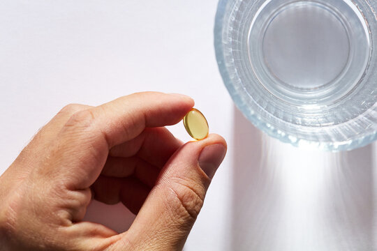 Person holding a vitamin softgel capsule next to a glass of water