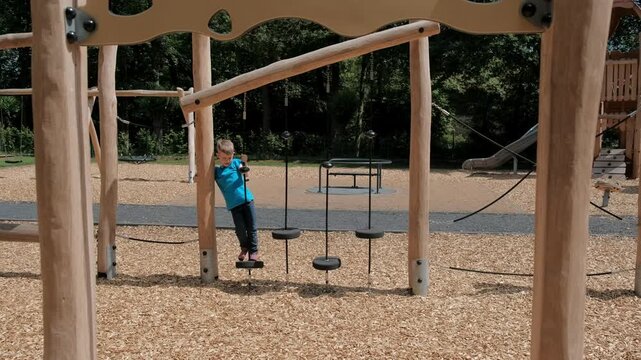 A focused young boy in a blue t-shirt carefully steps and balances on vertical wooden logs at a park obstacle course.