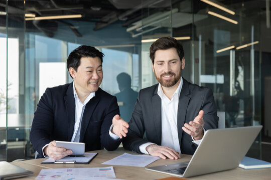 Two diverse male business partners collaborating and discussing a project while reviewing data on a laptop and tablet. Showing teamwork. Partnership. And professional communication in a modern office