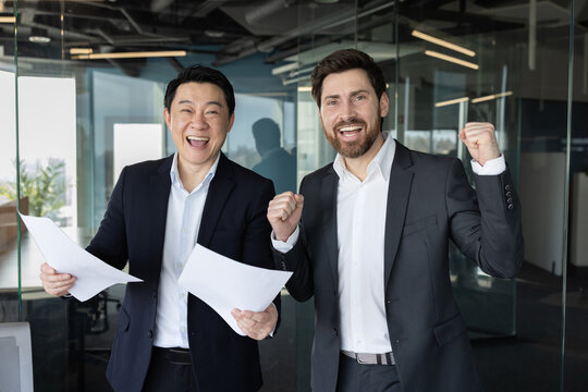 Two diverse business partners celebrating a successful deal, holding documents, expressing joy and achievement with cheerful faces and triumphant gestures in a modern office environment