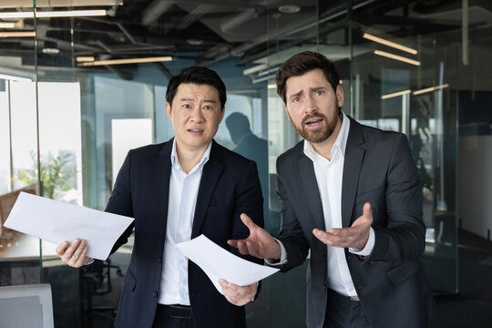Two diverse businessmen standing in a modern office, holding papers and expressing confusion or misunderstanding, portraying issues like miscommunication, stress, or problem solving