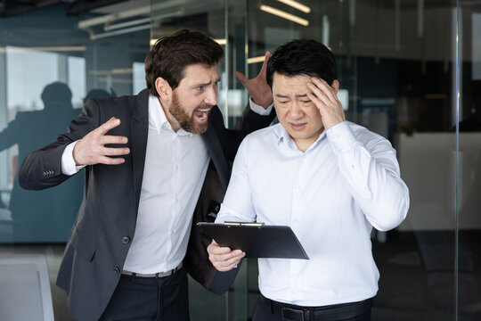 Aggressive male manager shouting at his frustrated asian male colleague holding a clipboard, symbolizing workplace conflict, harassment, and stress in a corporate environment