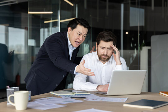 Angry asian boss is yelling and gesturing intensely at a stressed caucasian employee, who sits dejectedly at a laptop in a modern office, portraying workplace conflict and pressure