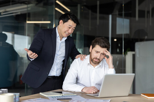 Asian male boss standing over a sad, stressed caucasian male worker at a laptop, showing workplace harassment and pressure in a modern office environment