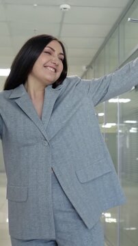 Cheerful female employee enjoying a dance of victory and success in the corporate corridor, expressing her happiness with energetic movements while holding a coffee cup and a blue folder