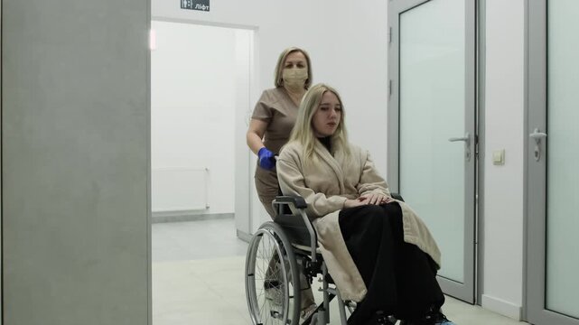 Nurse guides disabled woman toward sterile hospital room. Compassionate doctor supports patient moving through medical corridor. Slow motion