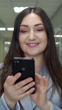 Cheerful female employee using a mobile phone to check her social media feed, chat with friends, or browse the internet during a break in a contemporary office corridor