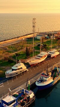 Yachts parked at quay in golden light, Nesebar, Bulgaria. Overhead drone view of large leisure vessels on stands, maintenance area, green park space and sea coast at sunset. Vertical video.