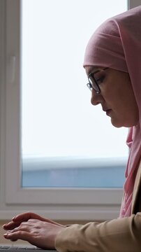 Focused muslim woman in a pink hijab and glasses typing on her laptop keyboard. Working diligently in an office environment with a window in the background. Showcasing modern islamic professionalism