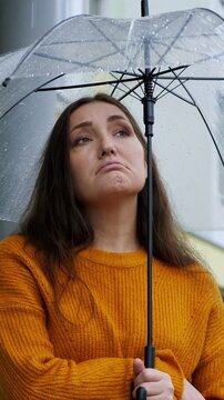 Sad young woman in a cozy sweater holding a transparent umbrella during an autumn downpour. Her melancholic expression conveys feelings of loneliness and thoughtfulness on a rainy day